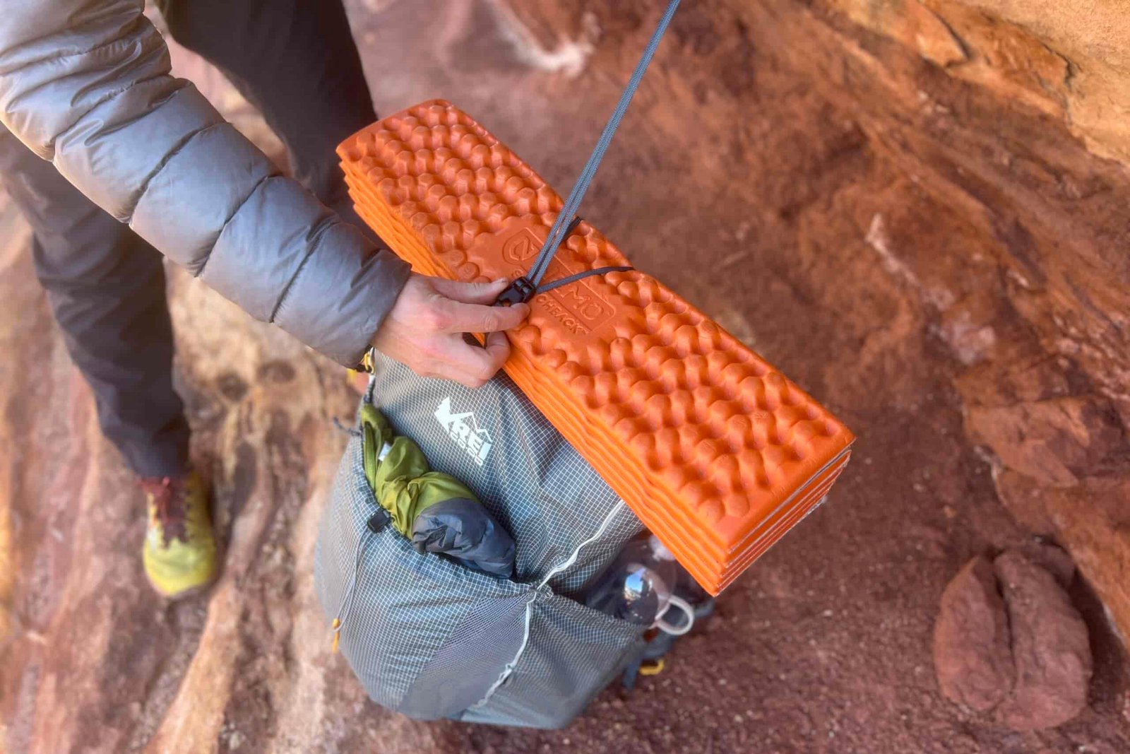 A hiker pulls on the top strap of a backpack with an orange sleeping pad on top and red rocks in the background.