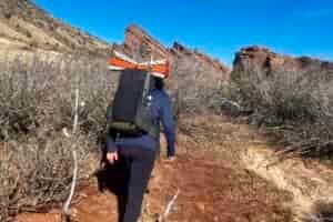 A hiker wearing a black backpack with an orange sleeping pad strapped to the top walks along a dirt trail, with red rock formations and scrubby desert vegetation visible in the background.
