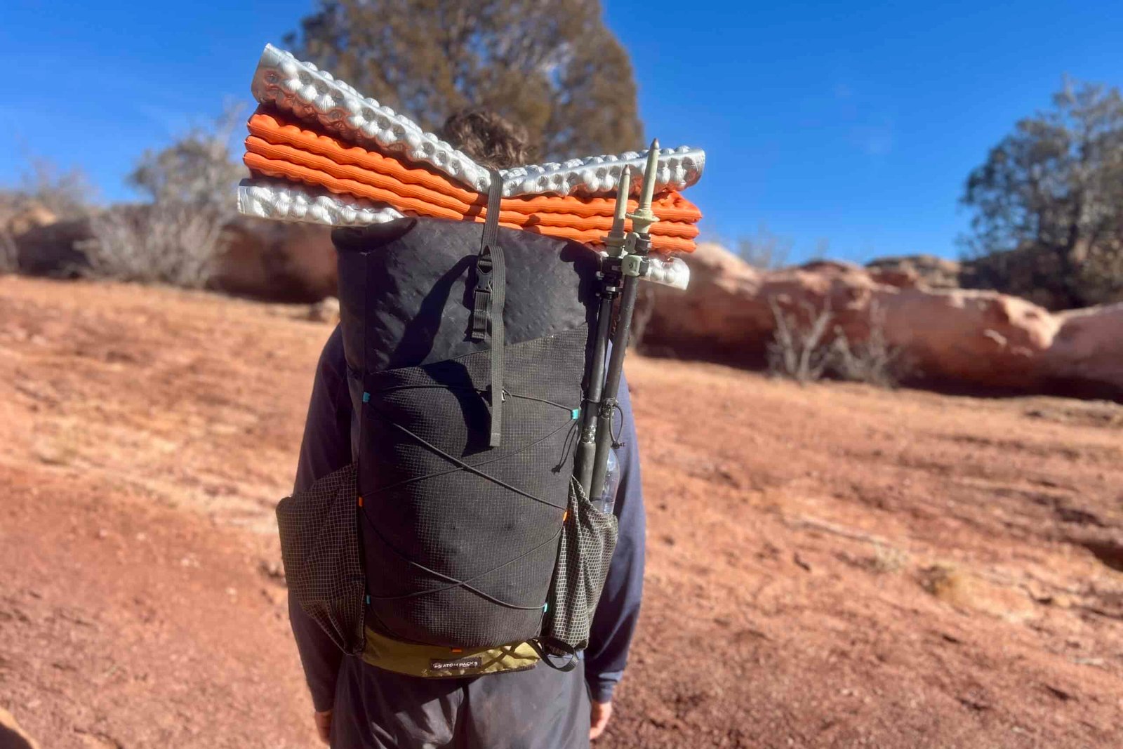 Back view of the Atom Packs backpack with orange and white sleeping pad secured on top and trekking poles mounted to the side.