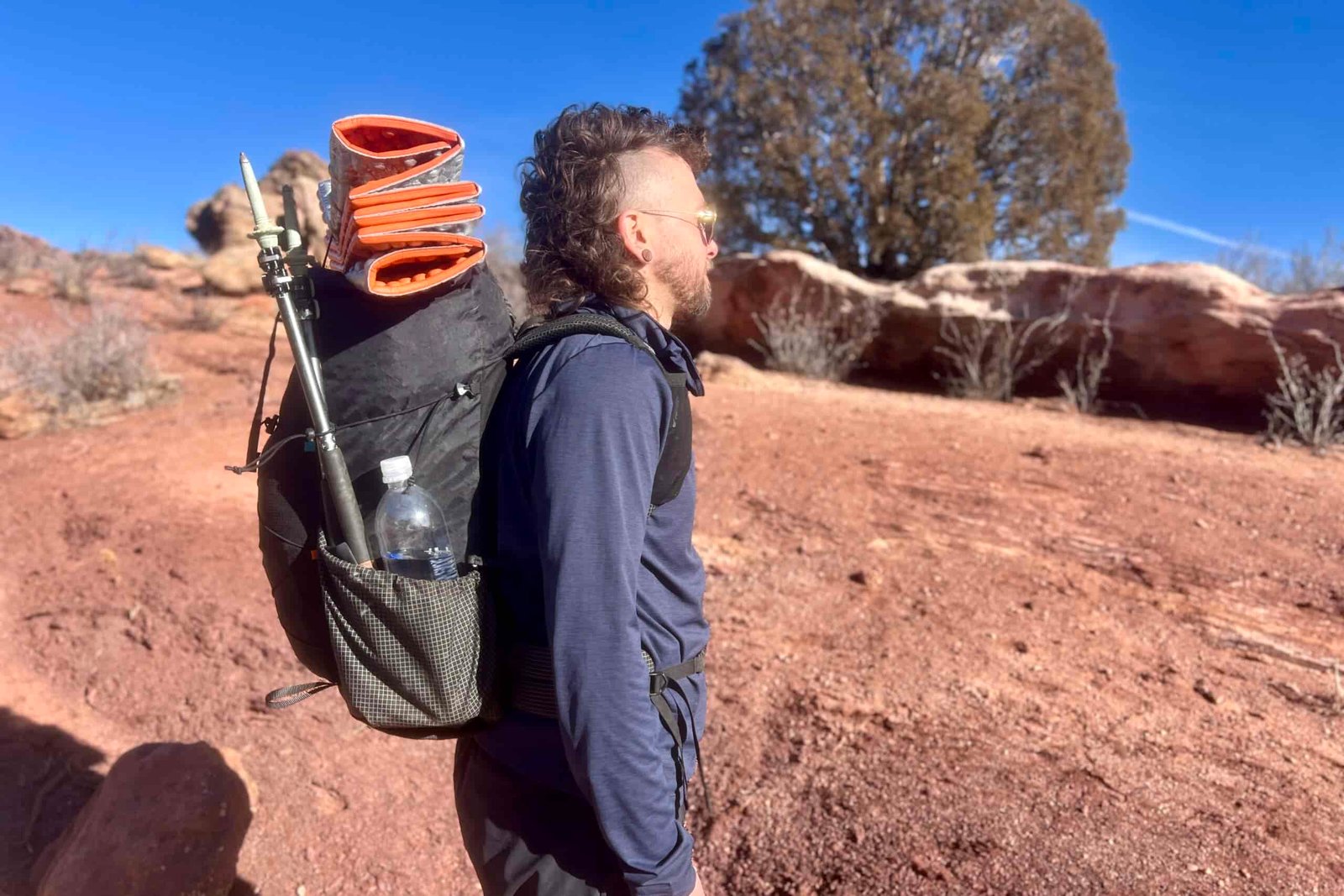 Profile view of a hiker wearing a blue long-sleeve shirt carrying the black Atom Packs backpack with sleeping pad and trekking poles attached, photographed against red desert rocks and clear blue sky.