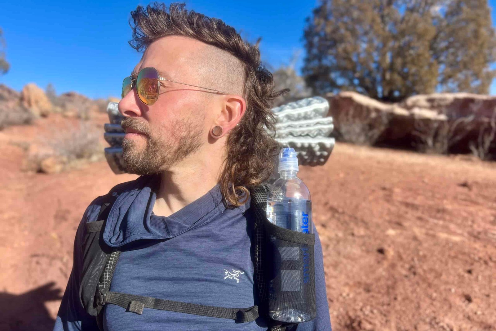 Profile view of a hiker wearing sunglasses with a water bottle secured in the pack's shoulder pocket, against a backdrop of desert terrain and blue sky.