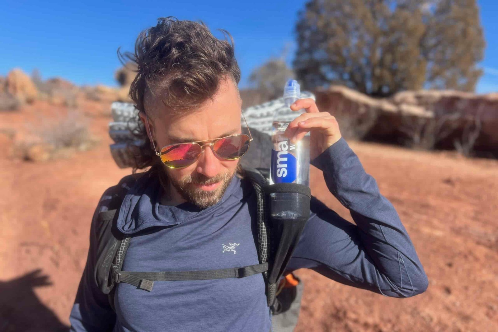 A hiker wearing mirrored sunglasses and reaching for a water bottle in the pack's shoulder strap pocket, with red rock formations visible in the background.