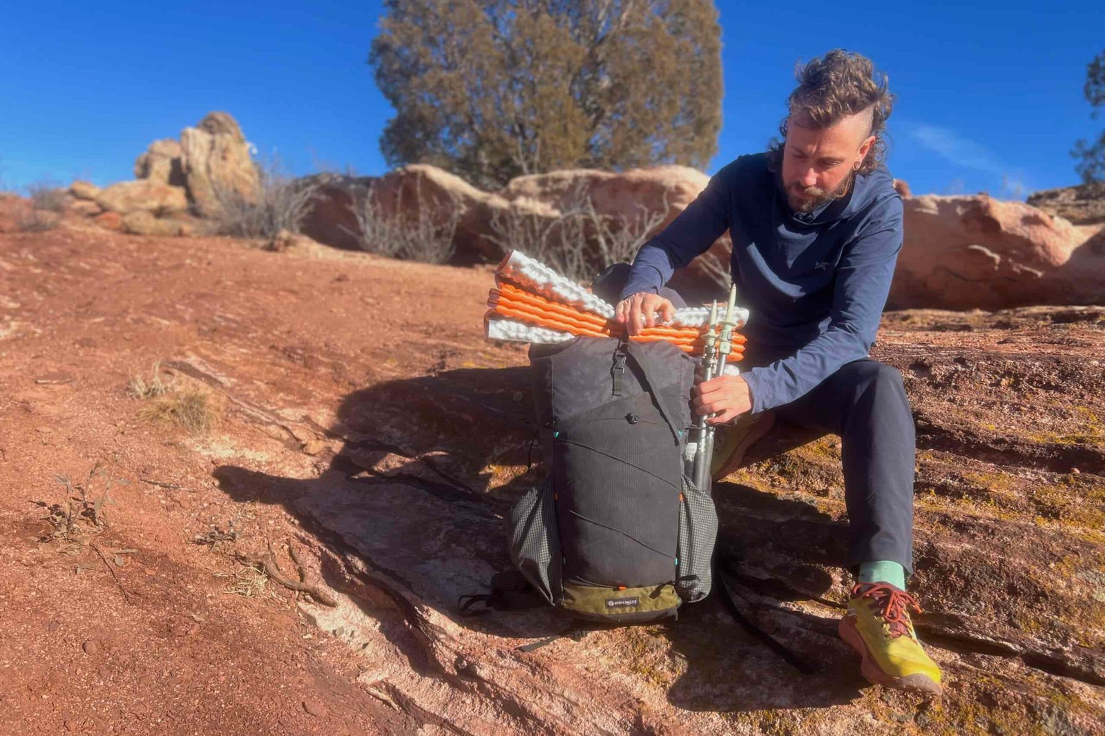 A person kneeling beside a black backpack on red desert rock, demonstrating how to attach an orange sleeping pad to the pack's top compression straps.