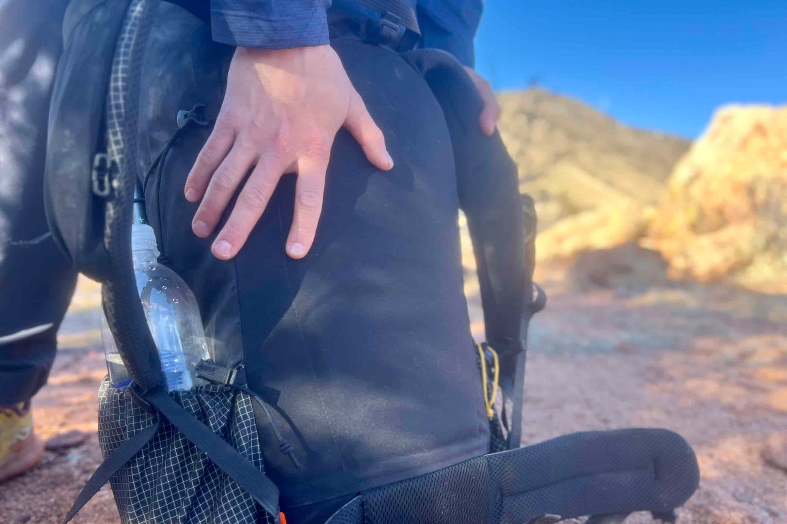 Close-up of a side pocket on a black backpack showing a water bottle storage solution, with desert mountains visible in the soft-focused background.