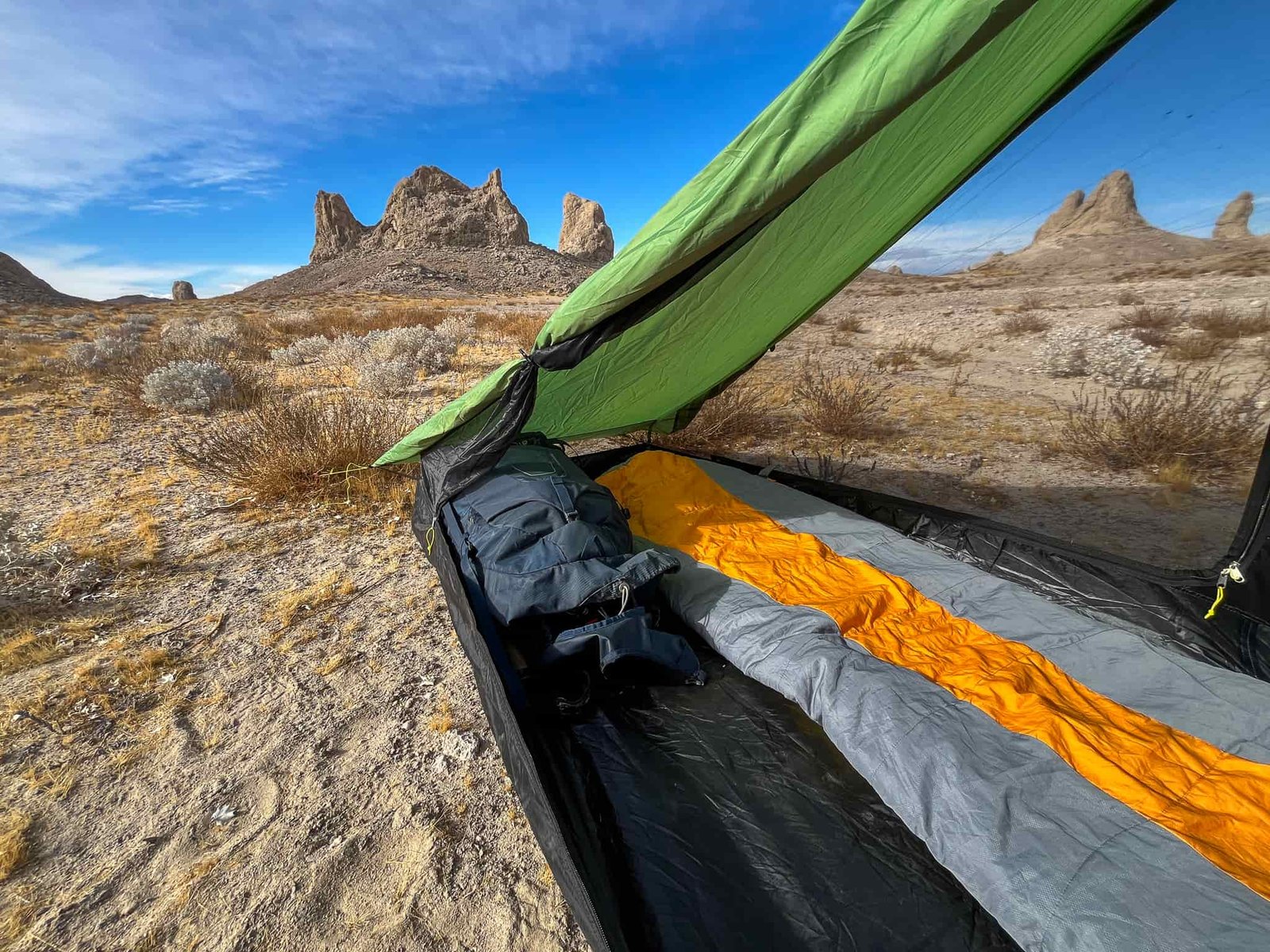 Interior shot of the Six Moon Designs Skyscape Scout. Shows a tied back door, backpacking pack, and sleeping bag inside. The blue sky and desert landscape visible in the background.