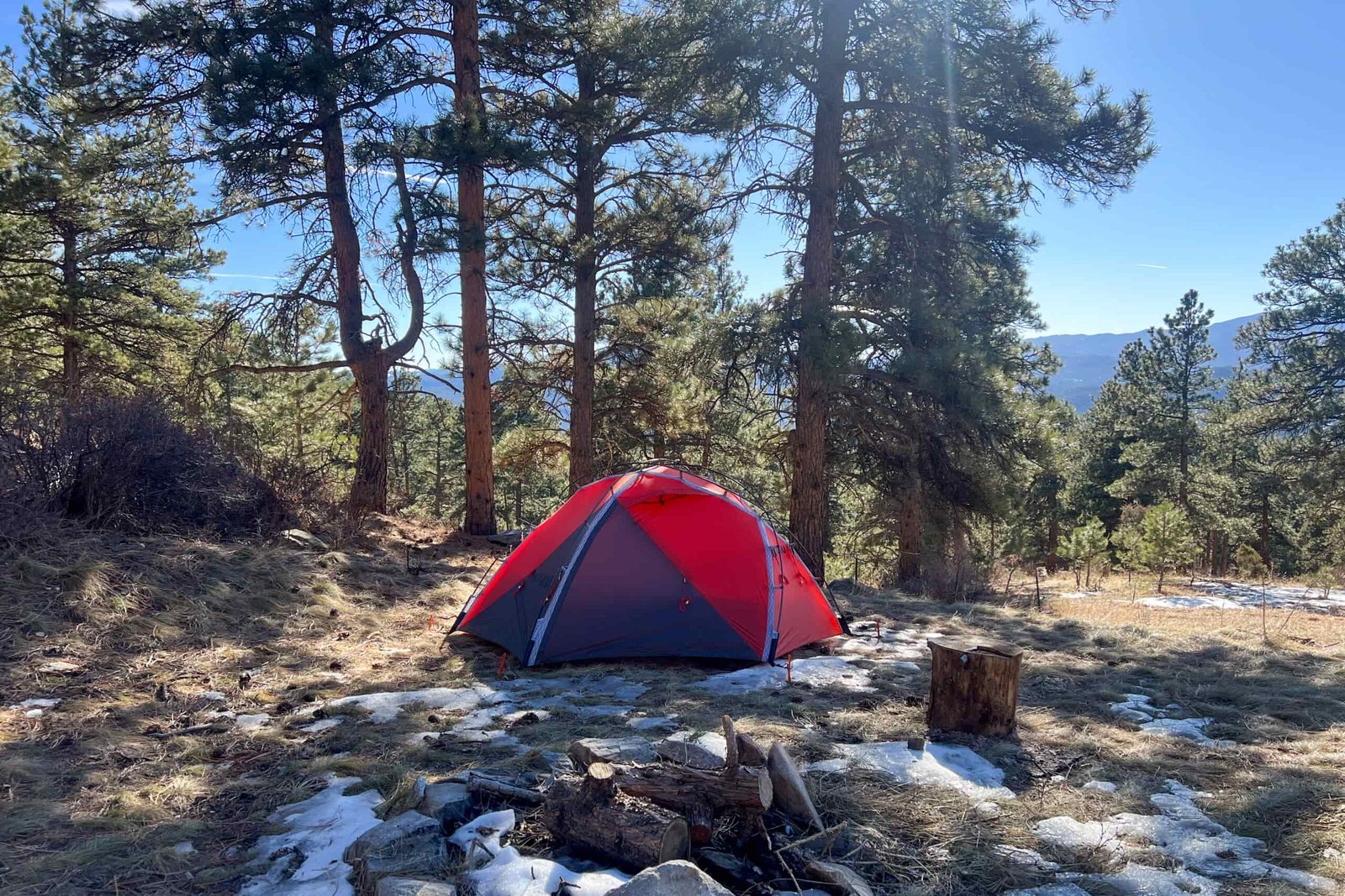 The KUIU Storm Star 2P tent set up in a forested campsite with a campfire ring nearby.