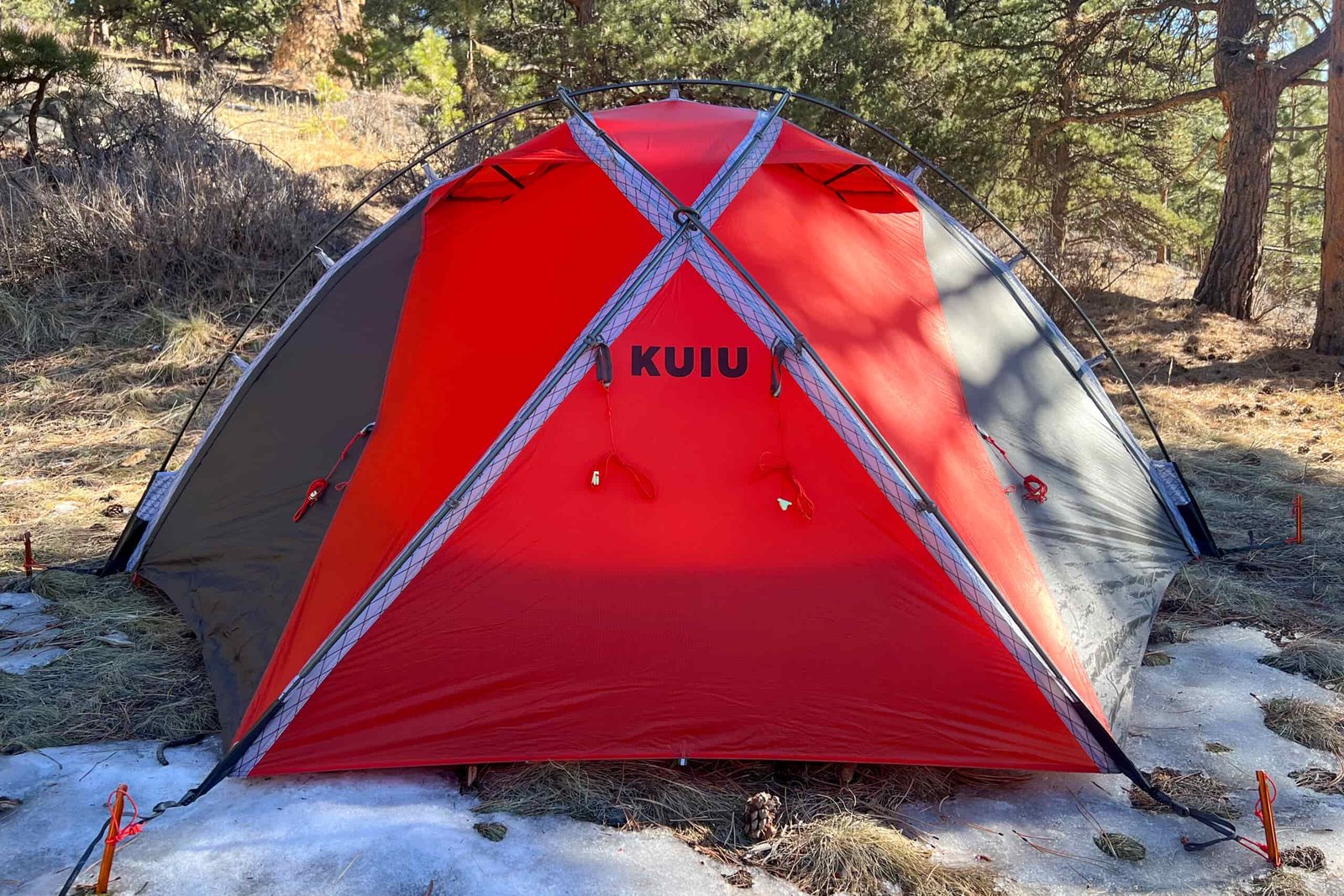 Front view of the KUIU Storm Star 2 tent, featuring its red and black color scheme and taut guylines on a forest floor.