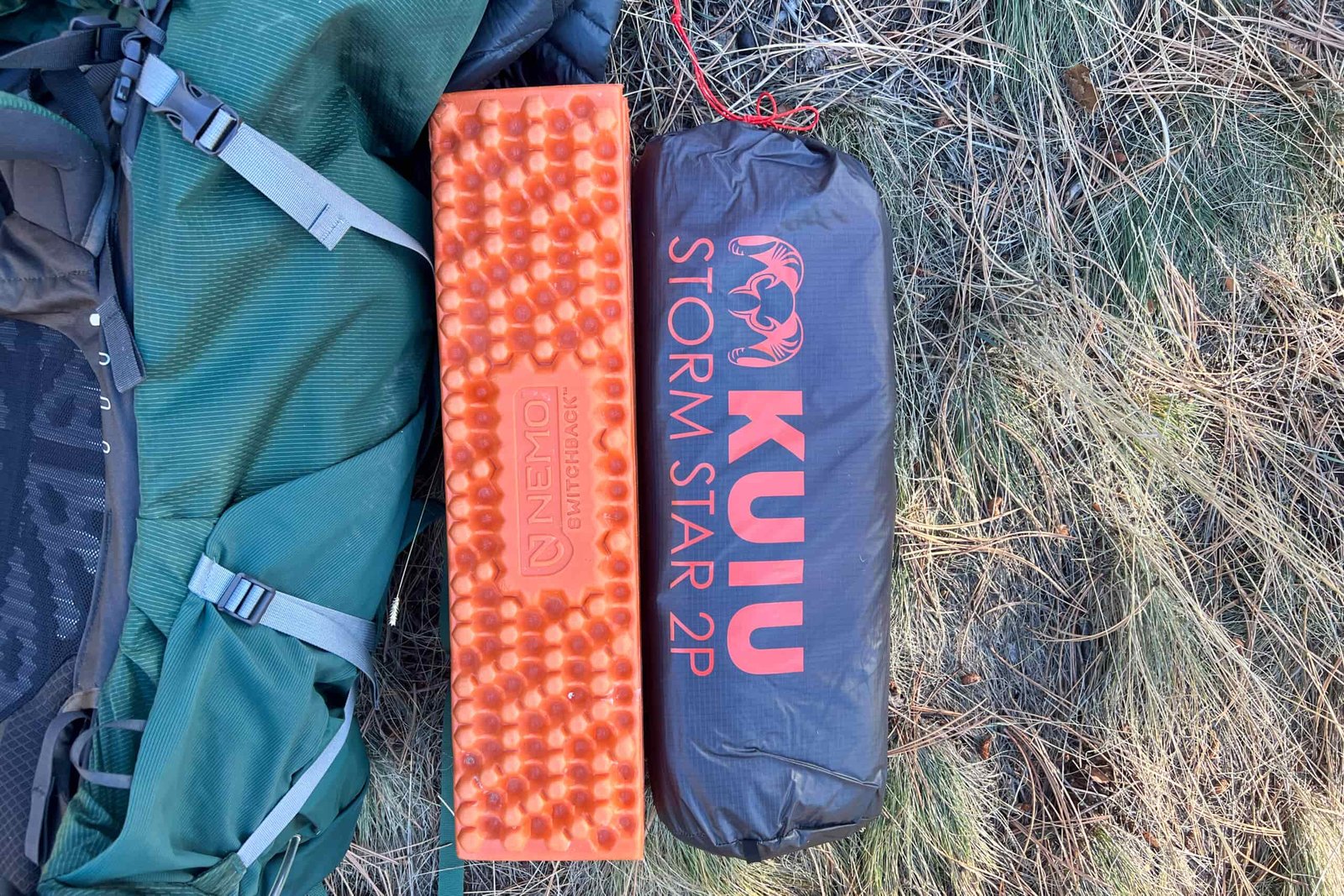 A packed KUIU Storm Star 2 tent bag next to an orange NEMO Switchback sleeping pad and a green backpack, ready for transport on the ground.