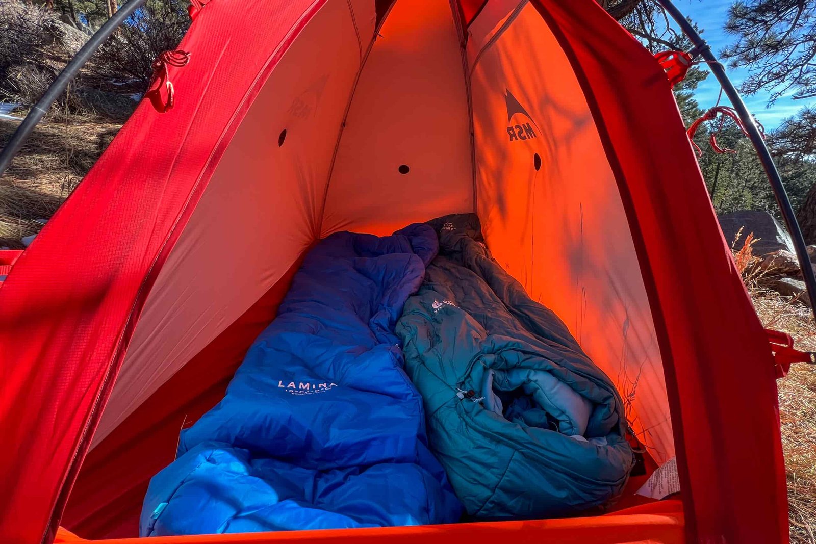 Inside the MSR Advance Pro 2 tent, two sleeping bags—one blue and one teal—are neatly arranged, with the bright orange walls reflecting sunlight.
