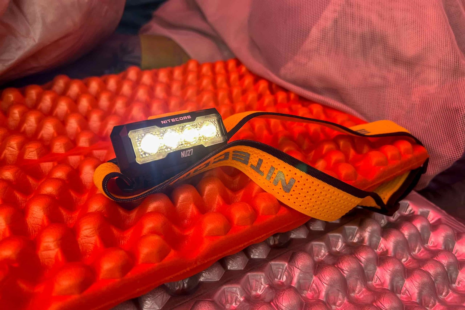 The photo features a headlamp sitting on a foam sleeping bad. The headlamp is on and pointed nead the camera.