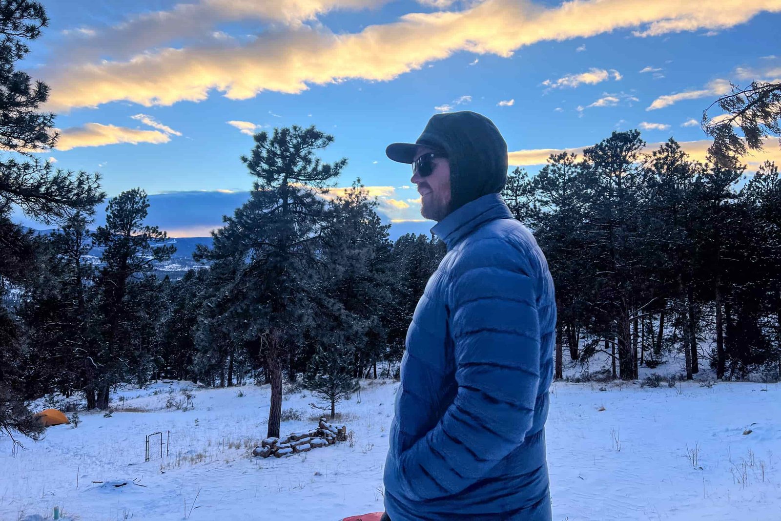 A man in a blue REI 650 Down Jacket standing in profile during a winter sunset, surrounded by snowy trees and a colorful sky.
