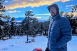 A man in a blue REI 650 Down Jacket stands with his hands in his pockets, gazing off into the snowy forest during an early winter evening.