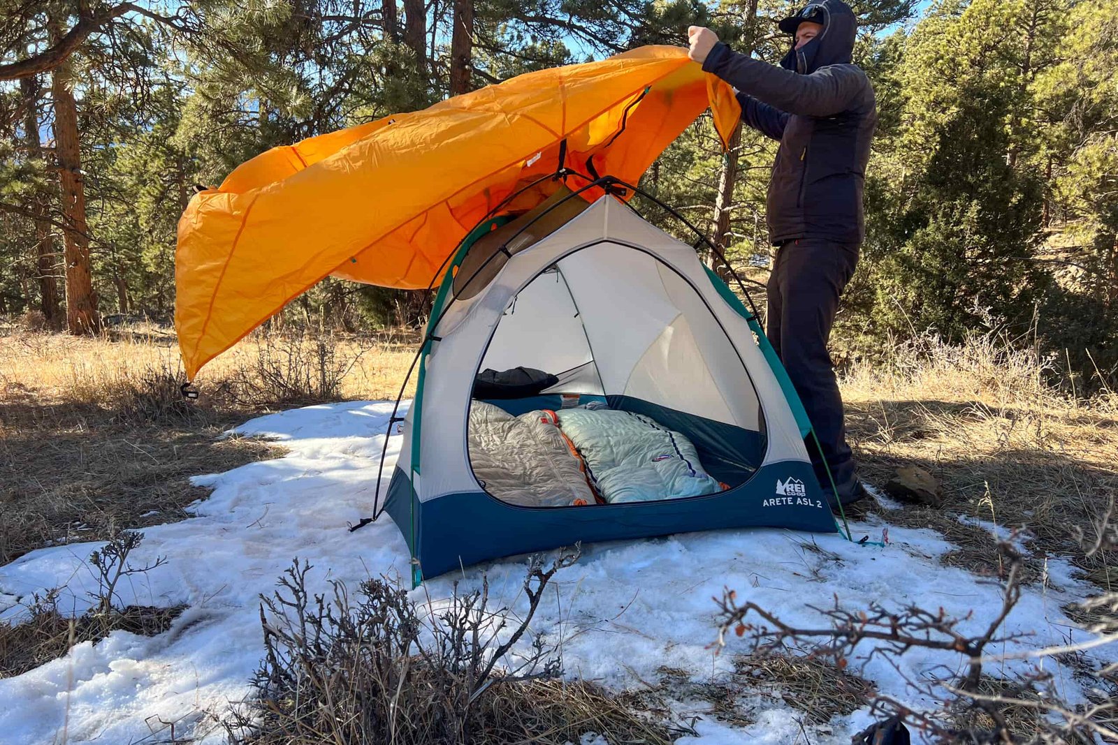 A man throwing the orange rain fly over a tent in a forested setting.