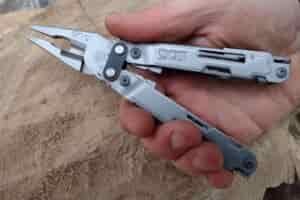 The silver SOG PowerAccess in hand, held above a log, with its needle nose pliers extended.