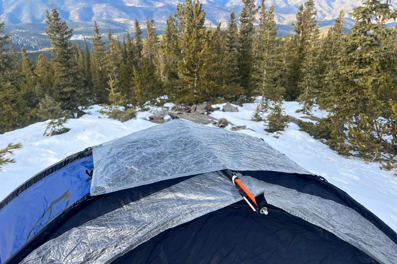 A close-up view of the Samaya 2.0 tent's reflective fly deployed over its structure, surrounded by snow-covered trees and distant mountain peaks.