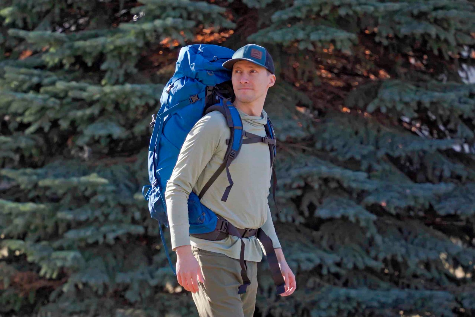 man wears blue backpack in front of a pine tree