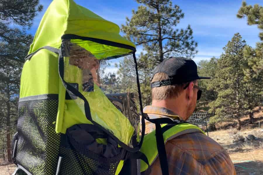 Rear view of a man hiking with a child in a bright green carrier featuring a transparent rain cover, surrounded by trees.