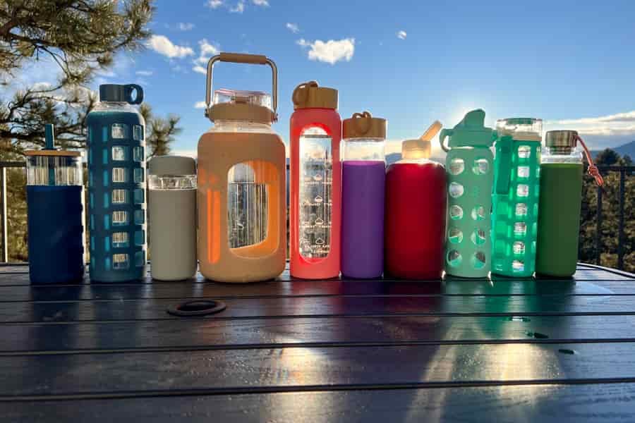 A variety of reusable water bottles arranged on a table with a scenic mountain sunset in the background, showcasing different shapes, colors, and designs.