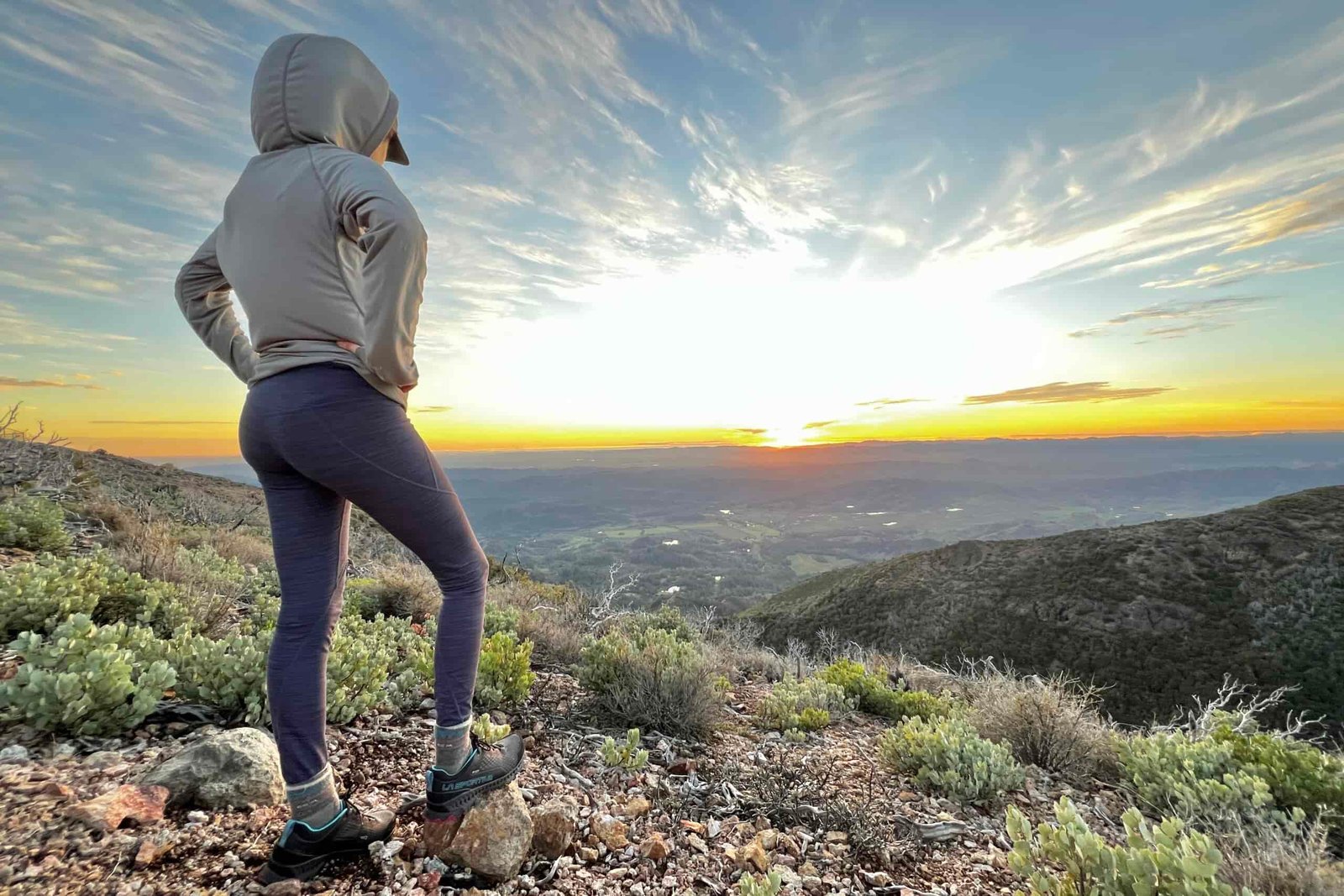 A woman stands before the setting sun with her hands on her hips.