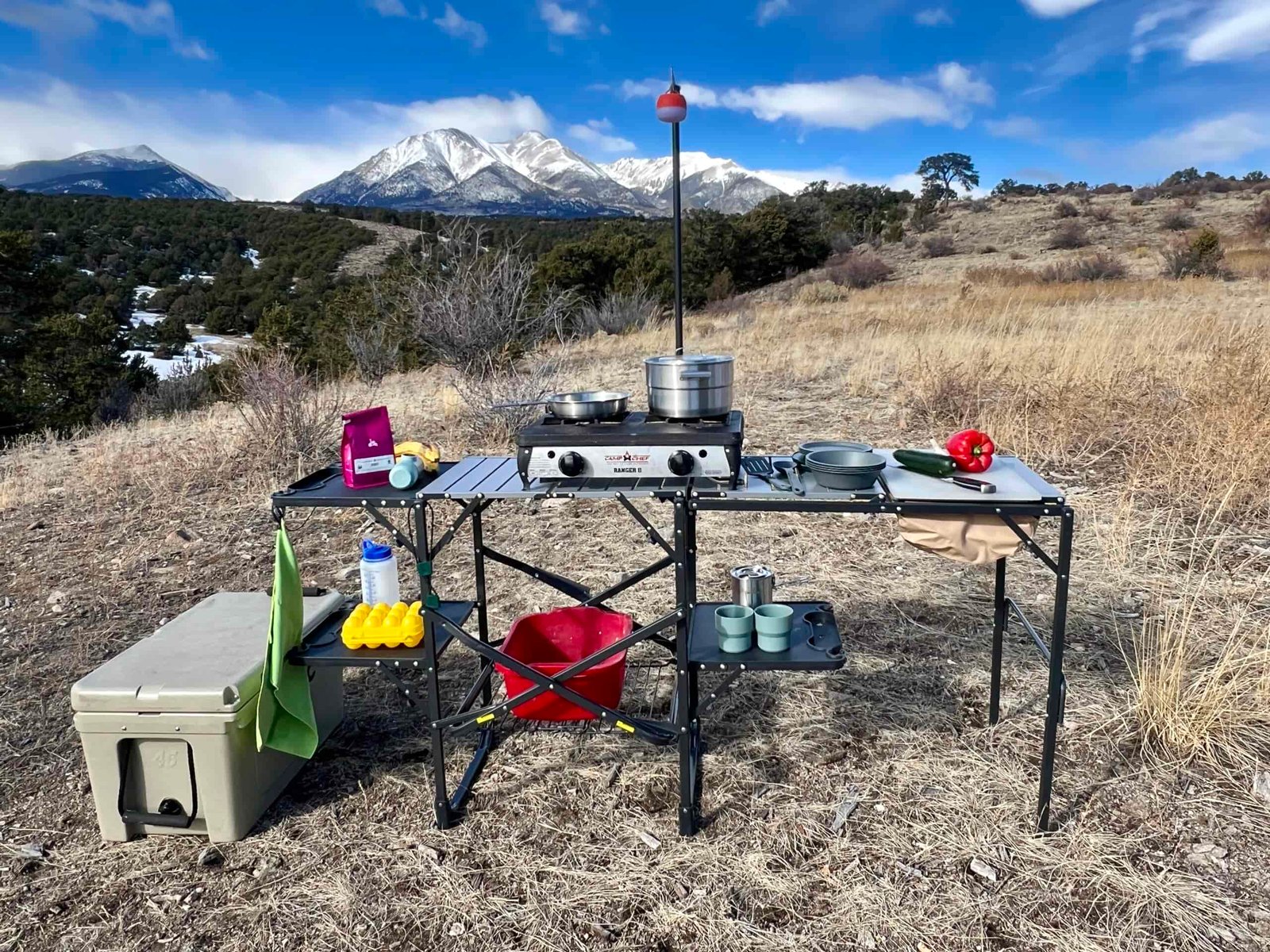 The GCI Master Cook Station stands outside with a mountain backdrop. Its shelves hold kitchen items and an outdoor stove. A cooler is on the ground next to it.