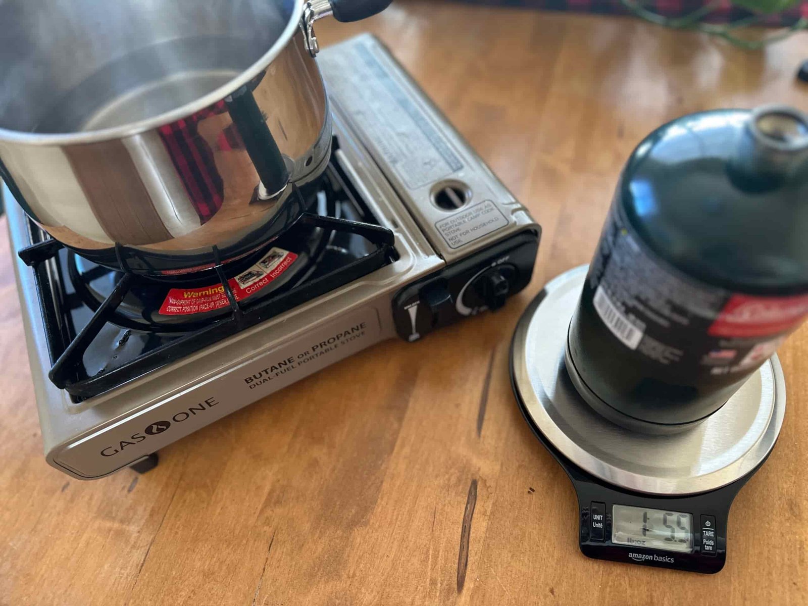 a scale with a propane canister on top with a camp stove in the background to test the amount of fuel used to boil water.