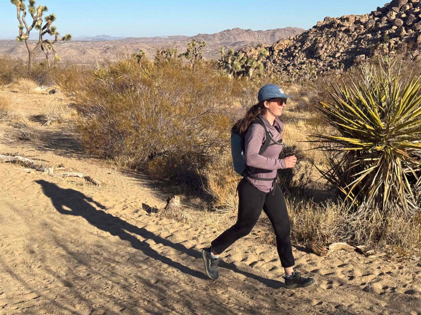 A runner runs down a sandy trail with a light day pack. The day pack has a hydration hose running out of it, and the hose is secured to the sternum strap. In the background is a desert scene with low dry mountains and Joshua Trees.