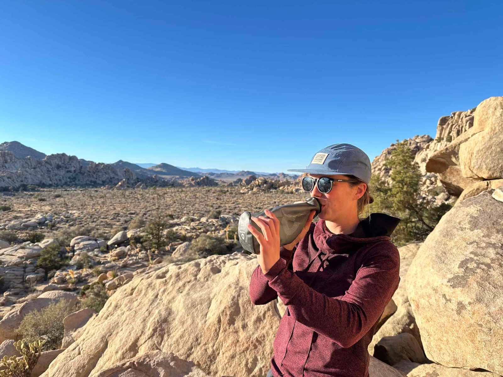 A person wearing sunglasses and a sun hat drinks from the HydraPak seeker water bag. The background is of Joshua Tree National Park, and it shows granite rock formations and some trees.