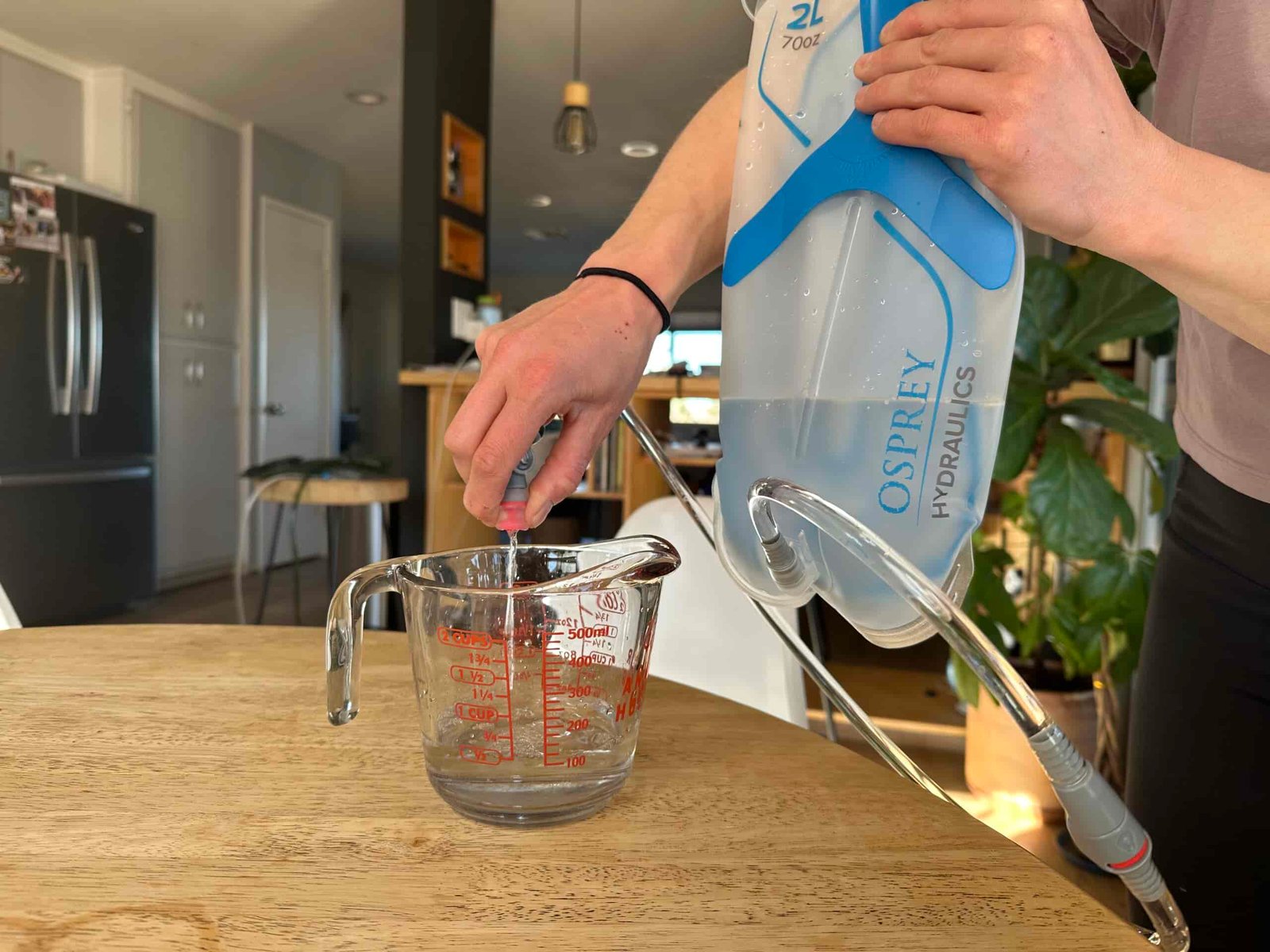 Close up photo of a person standing in a kitchen and holding the Osprey Hydraulics bladder in the air while squeezing the bite valve, allowing water to flow into a measuring cup.