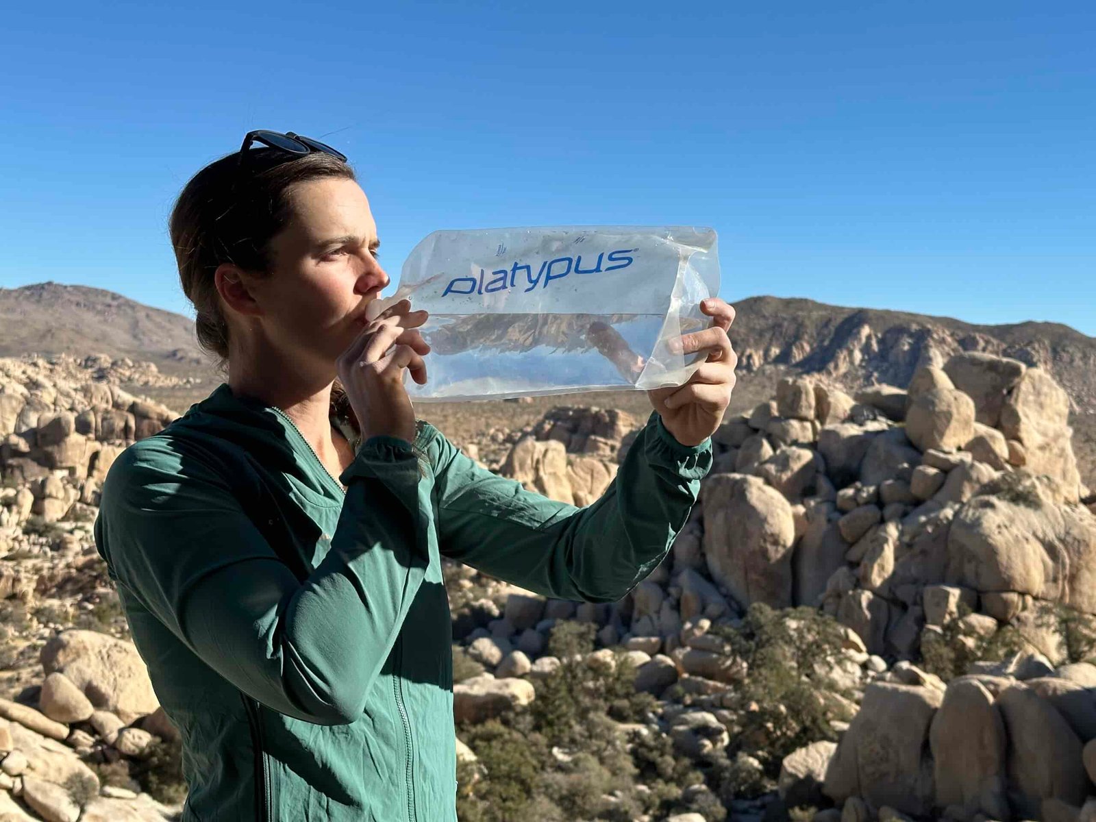 A person drinks out of the Platypus Platy, holding it with two hands. In the background are large granite rock formations.