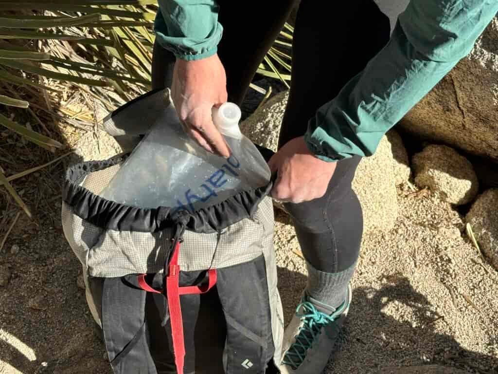 Close-up of a the Platy being loaded into the top of a small climbing pack. The climber is standing in the sand with a granite boulder and desert plant behind her.