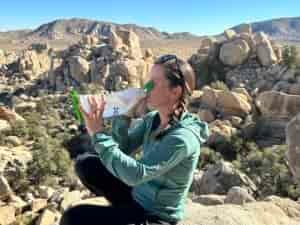 A hiker drinks from the CNOC VectoX water container, using two hands to hold it. The granite rock formations of Joshua Tree National Park are in the background.