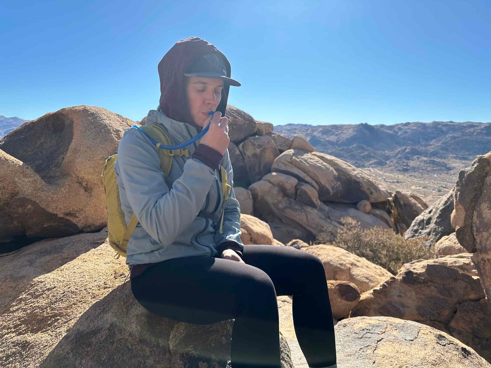 A hiker sips out of the CamelBak Crux's hydration hose while sitting on a granite boulder. In the background are low, dry mountains of the Southern California desert