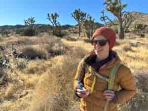 A hiker smiles on a trail with Joshua Trees in the background, holding the bite valve of the Platypus Hoser Reservoir.