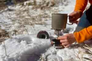 A close up of a hiker holding the evernew pasta pot on a stove with one hand and turning the stove on with the other. The lid of the pot is sticking out of snow.