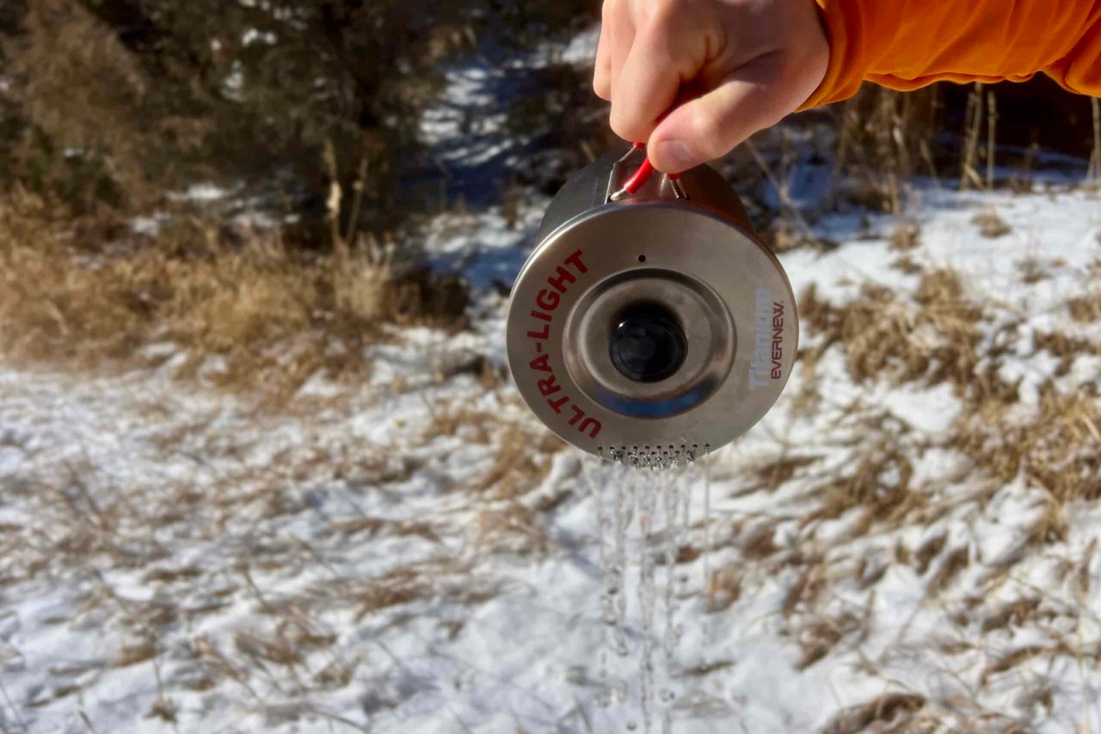 Close-up view of the Evernew Pasta Pot's titanium lid showing the distinctive pattern of drain holes and 'ULTRA-LIGHT' text engraved on the metallic surface while a hiker pours water through the holes. Photographed against a snowy winter background.