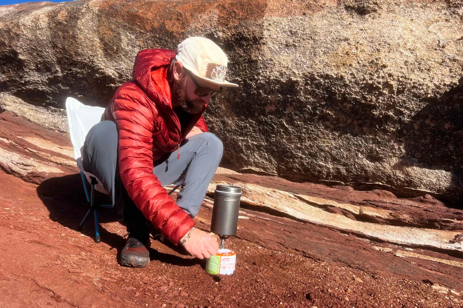 A hiker sits in a chair and lights a backpacking stove with a pot on top against a red rocks background.