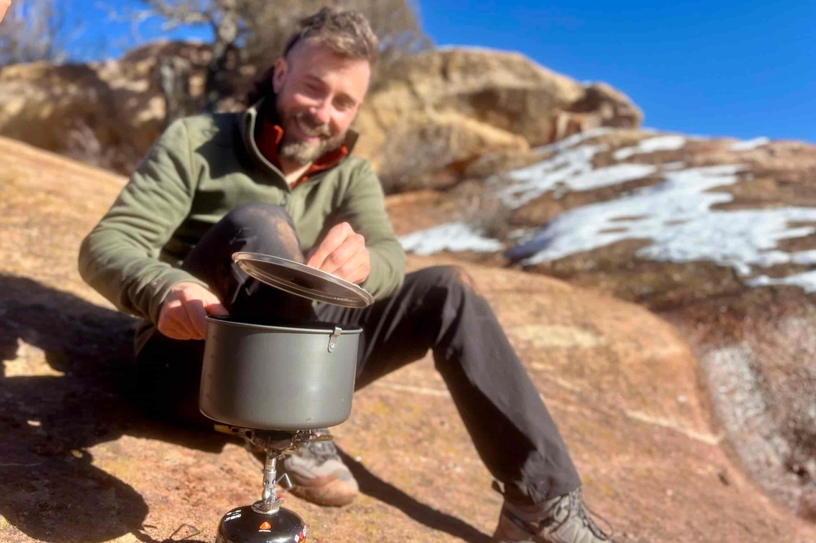 A hiker sits on the ground and lifts a pot's lid while cooking with it on a backpacking stove. Snowy desert background.
