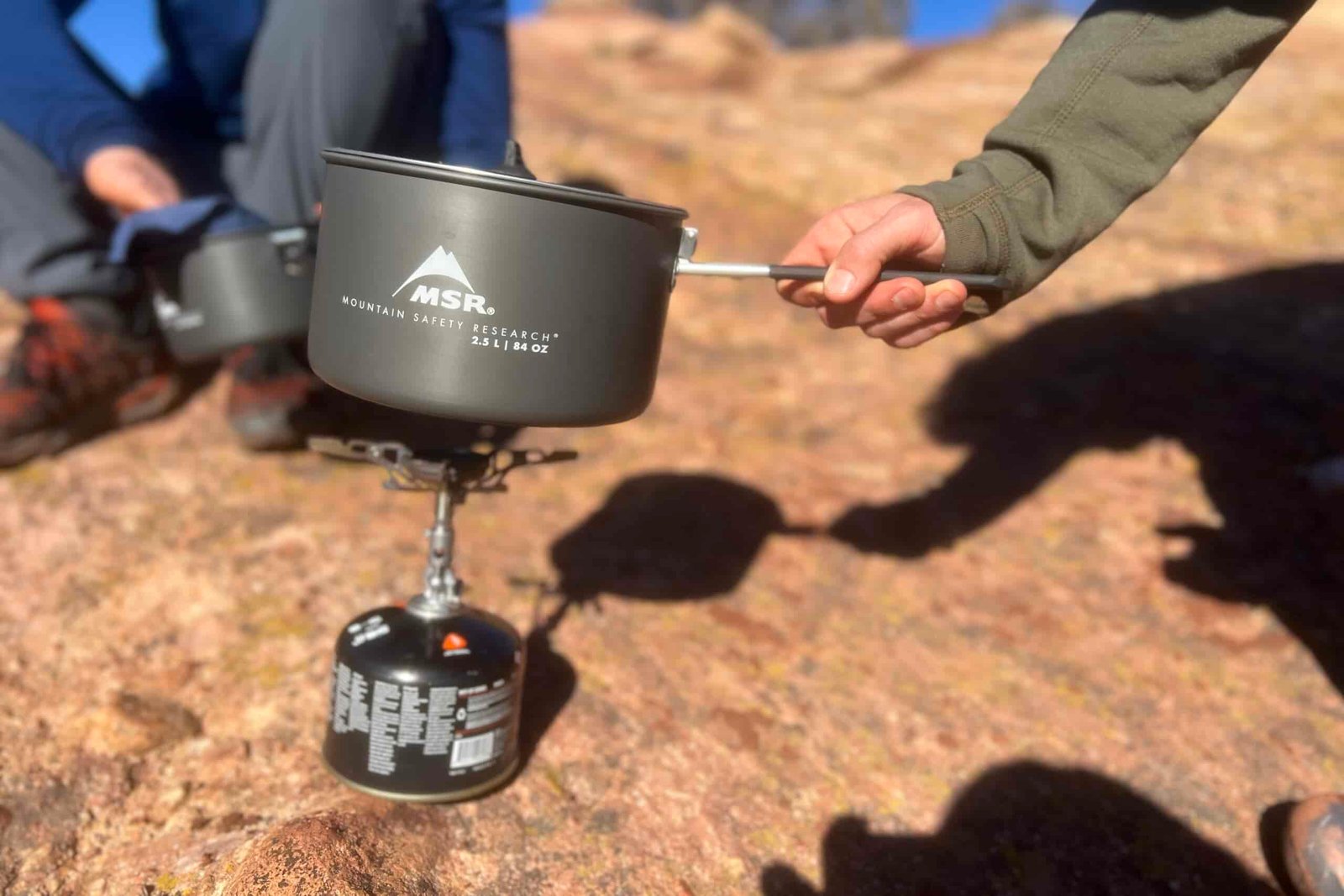 A hand places a pot on a backpacking stove while a hiker washes a pot in the background. Set in a rocky desert setting.