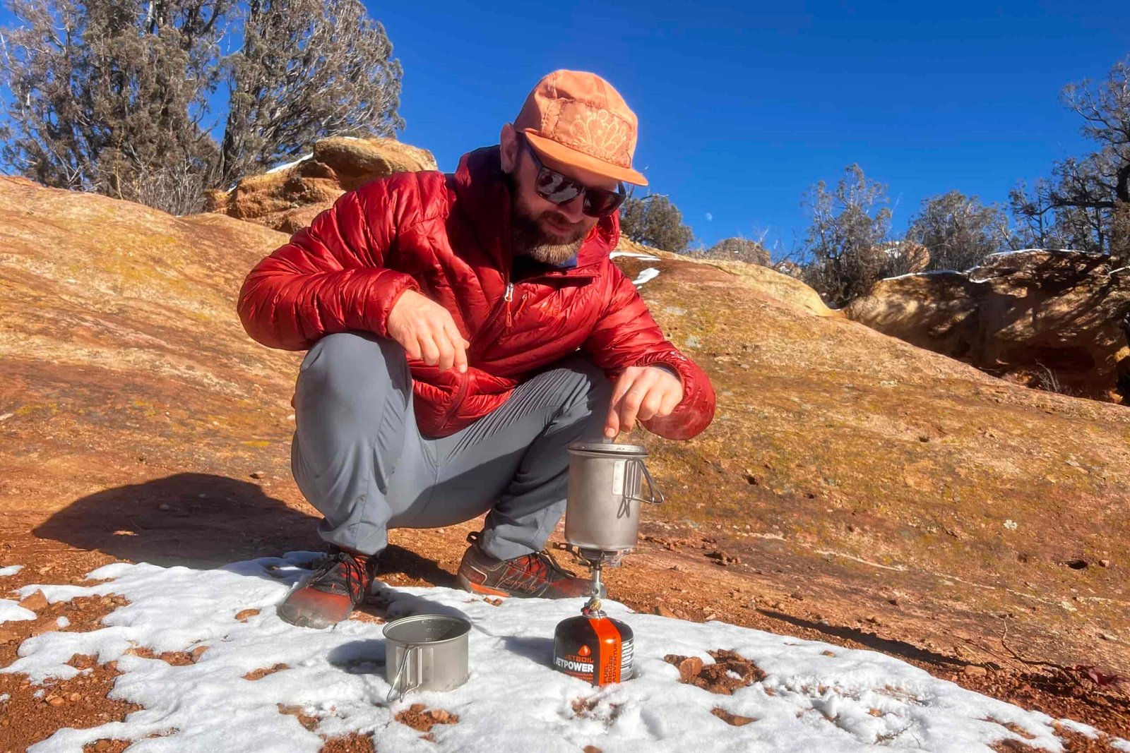 A hiker squats next to a cook pot and lifts the lid. Red rocks and desert are in the background.