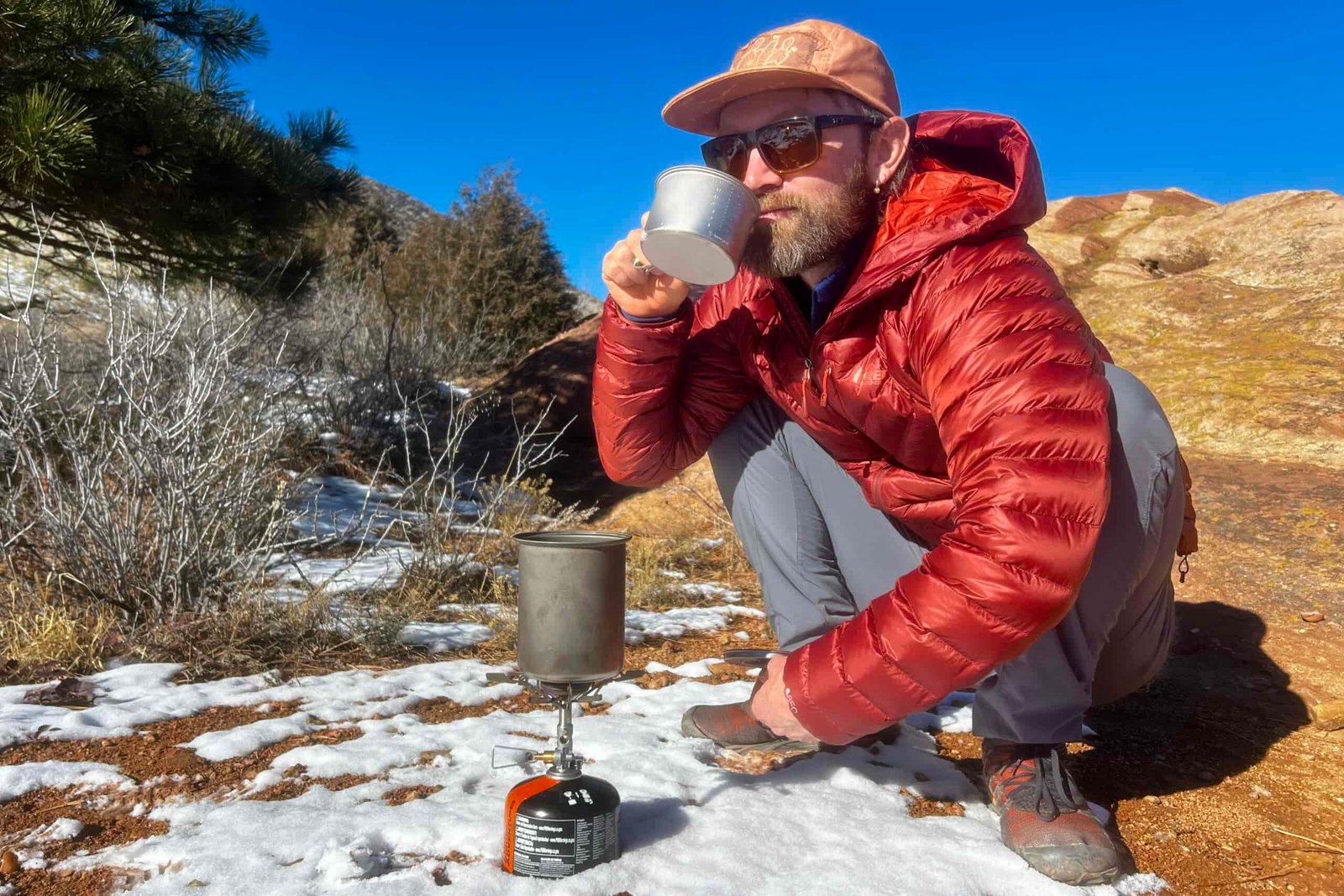 A hiker squats next to a camping cook set and sips from a small cup against a desert background.