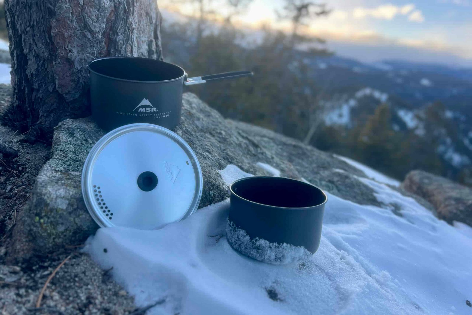 Two cook pots and a lid sit next to a tree with snow on the ground and a mountain sunset view.