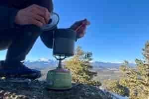 A hiker crouches and cooks with the trail mini solo on a backpacking stove with a mountainous background.