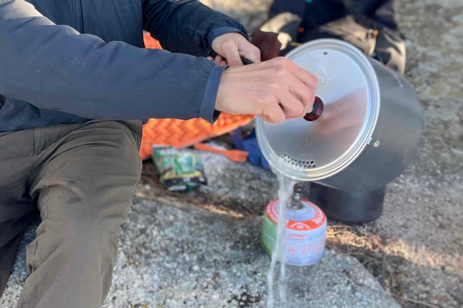 A hiker strains water out of a pot with backpacking gear strewn in the background.