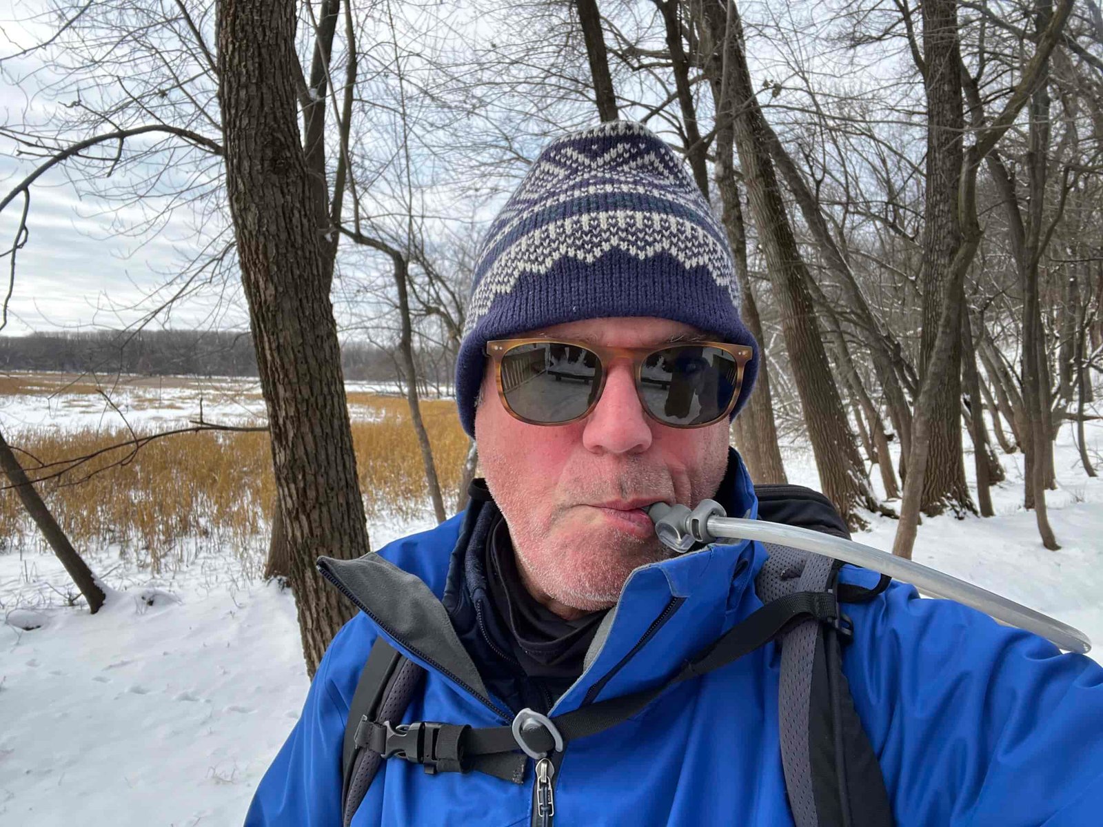 Close up of a snowshoer with a winter hat and sunglasses on. The background is snowy and lightly forested. The snowshoer is drinking from the HydraPak Contour's bite valve.