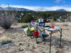 The GCI Master Cook Station stands outside with a mountain backdrop. Its shelves hold kitchen items and an outdoor stove. A cooler is on the ground next to it.