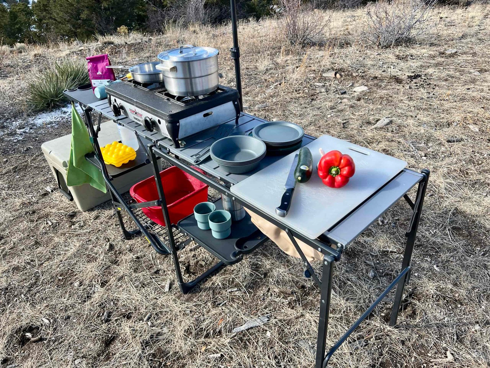 The GCI Master Cook Station stands outside in a scrub and desert setting. Its shelves hold kitchen items and an outdoor stove, and a cooler is on the ground next to it.