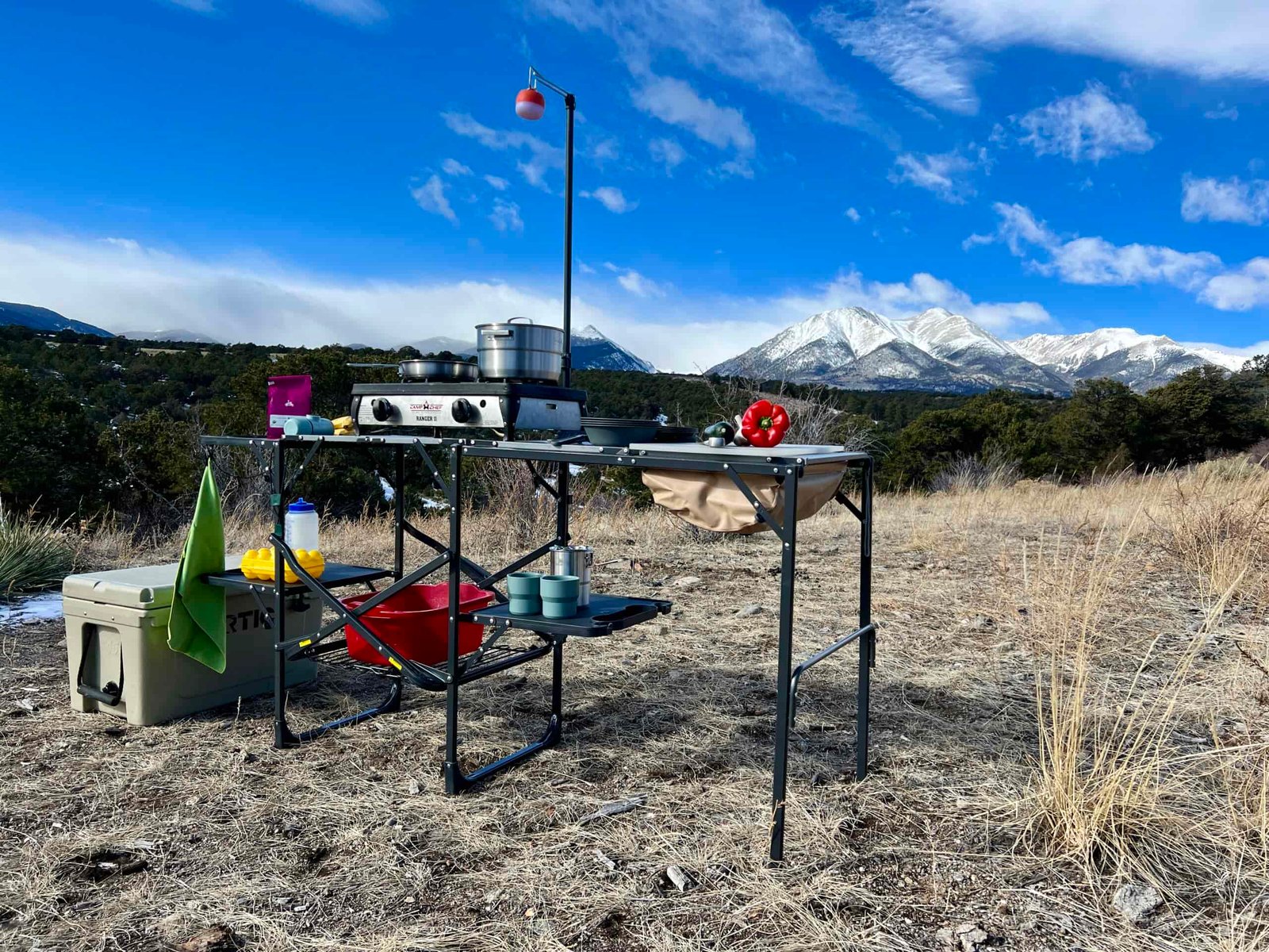 The GCI Master Cook Station stands outside with a mountain backdrop. Its shelves hold kitchen items and an outdoor stove. A cooler is on the ground next to it.