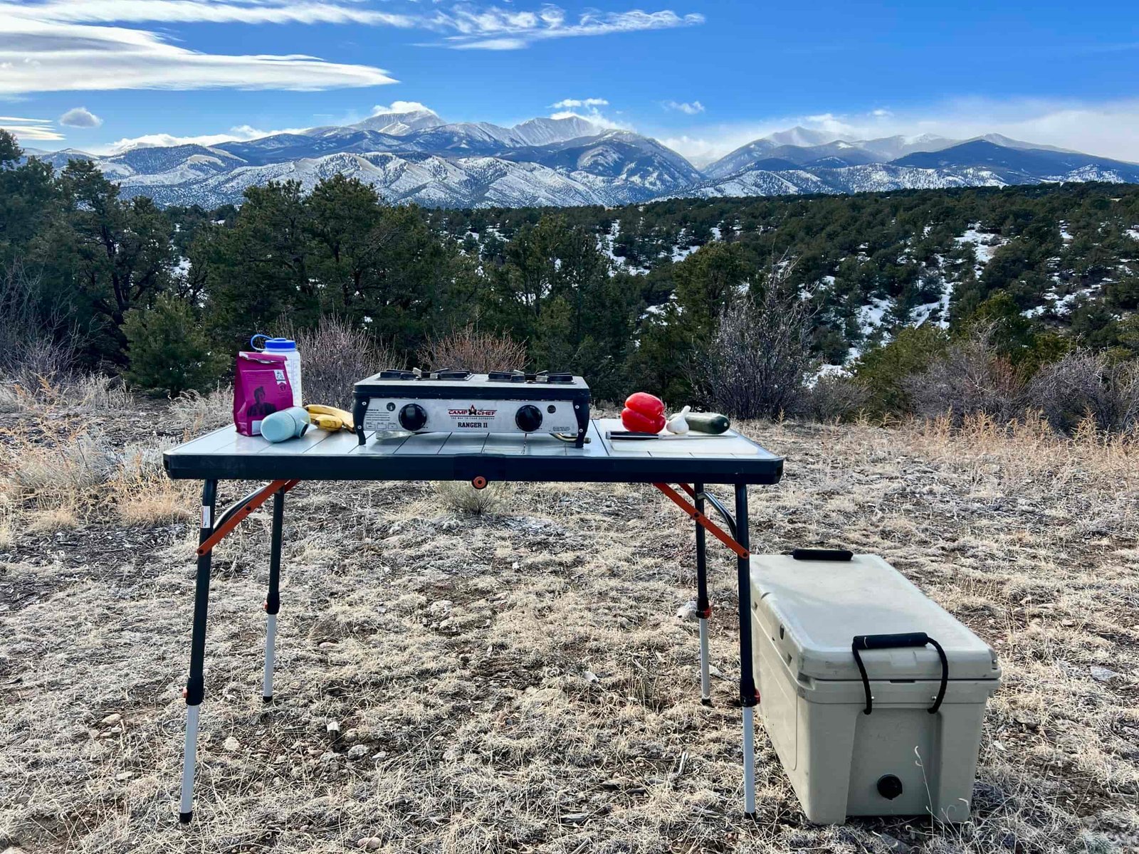 The REI Camp Prep Table has a stove, a red pepper and zucchini, a pink coffee bag, and bananas.