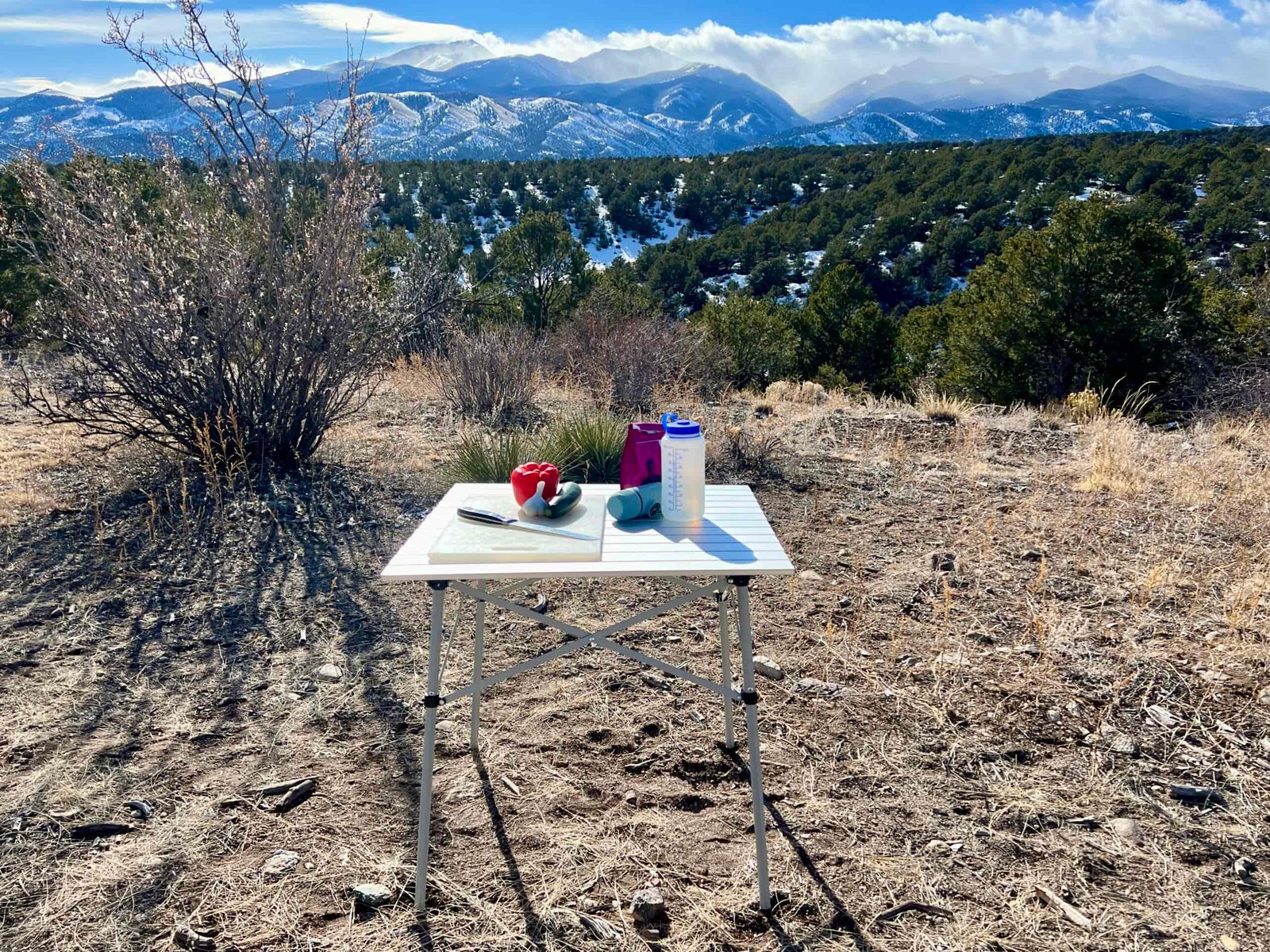 The Coleman Compact Roll Table stands on dirt and scrub with a scenic mountain backdrop. A Nalgene, cutting board, knife, and veggies are on top.
