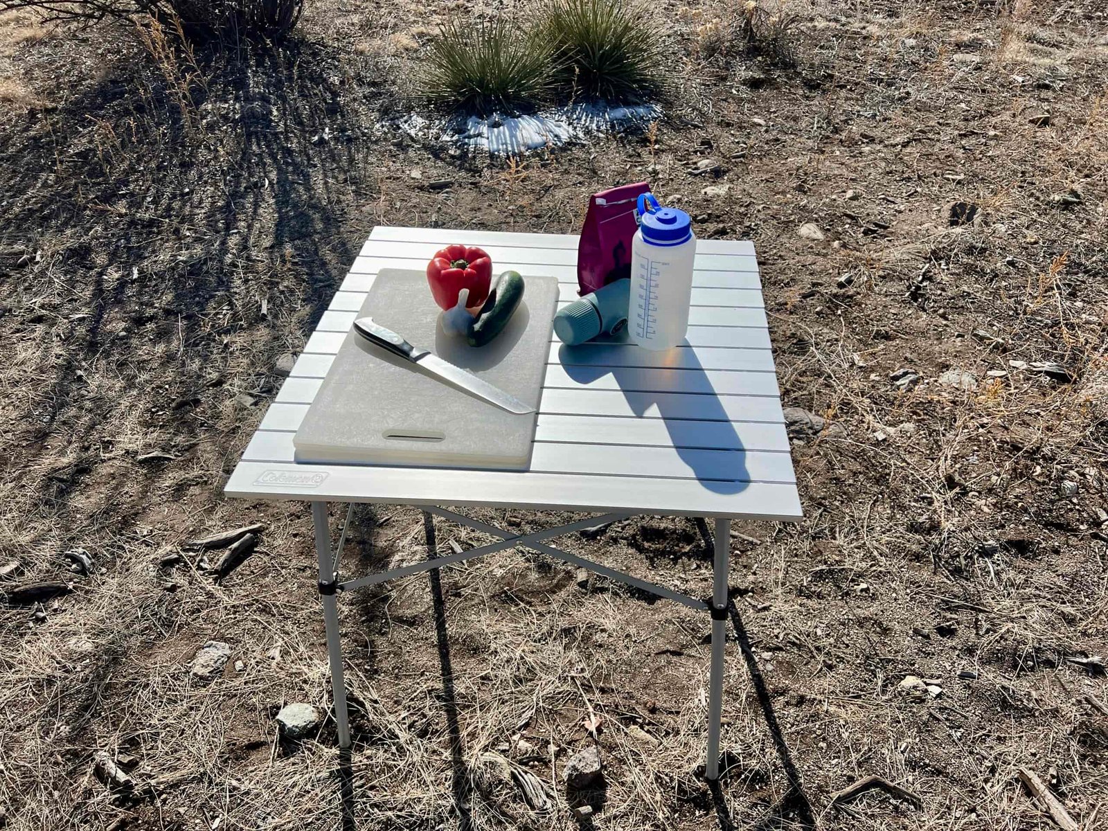 The Coleman Compact Roll-Top Table stands on dirt and has a cutting board, red pepper, zucchini, garlic, a Nalgene bottle, coffee, and Aeropress on its top.